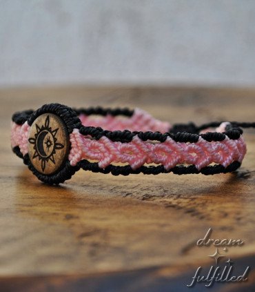 hand holding a black and pink braided thread bracelet and laser-engraved wood with a sun, moon, and star symbol