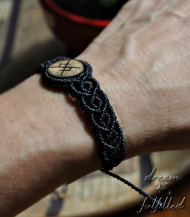 Side view of wrist with a wooden bracelet and black macramé thread, engraved with a bindrune symbolizing eternal love.